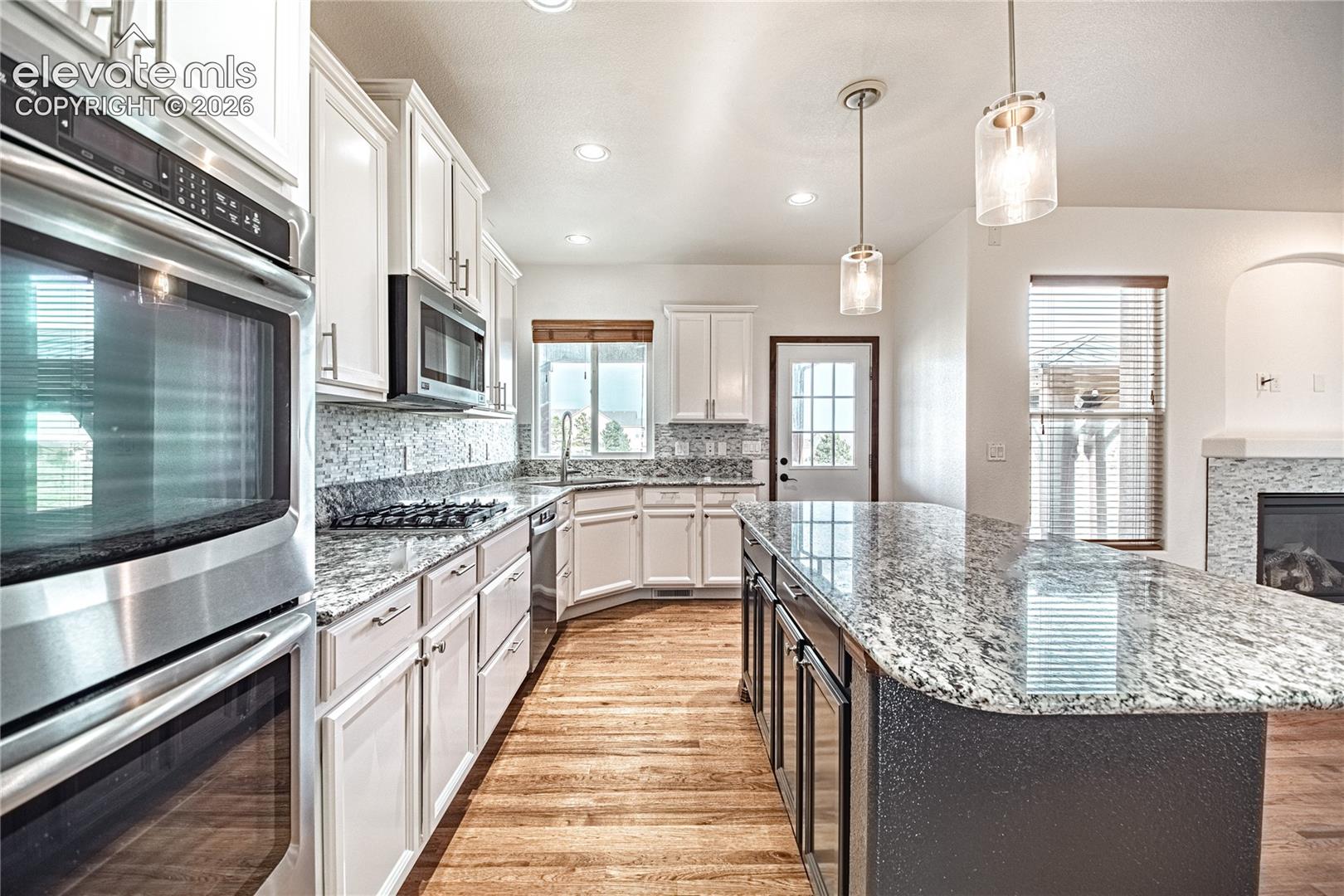 Spacious kitchen with a large central island featuring a stone countertop, white cabinetry, integrated stainless steel appliances, wood-finish flooring, and pendant lighting