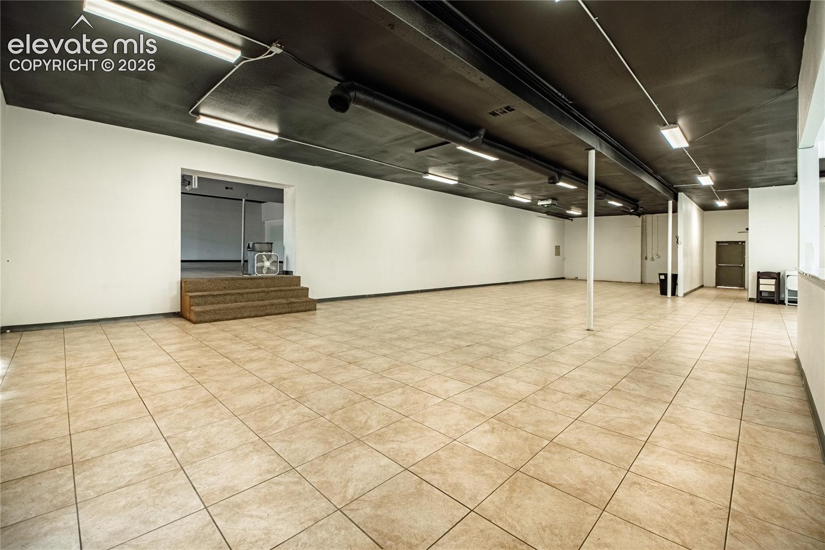 Expansive open-concept space featuring neutral-toned tile flooring, white walls, and a dark-painted ceiling with exposed conduit