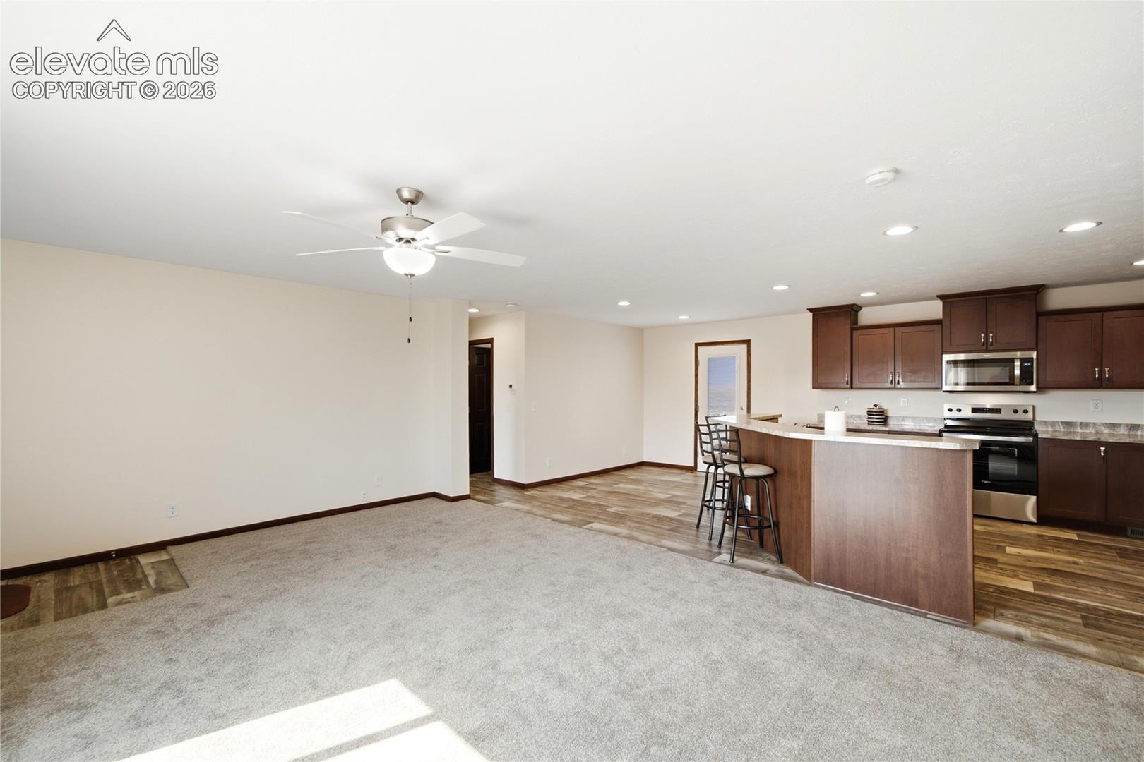 Open-concept living and kitchen area featuring recessed ceiling lighting, ceiling fan, wood-finish flooring, rich wood cabinetry with stainless steel appliances, and a central island with seating