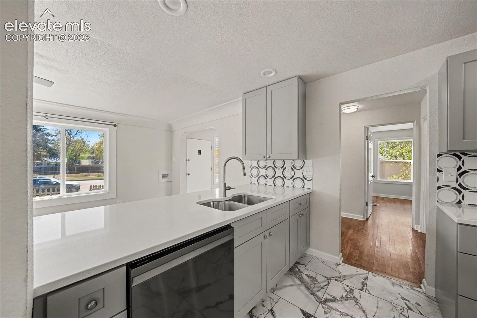 Kitchen featuring grey shaker cabinetry, quartz countertops, a dual-basin stainless steel undermount sink with a high-arch faucet, patterned tile backsplash, and large-format veined floor tile