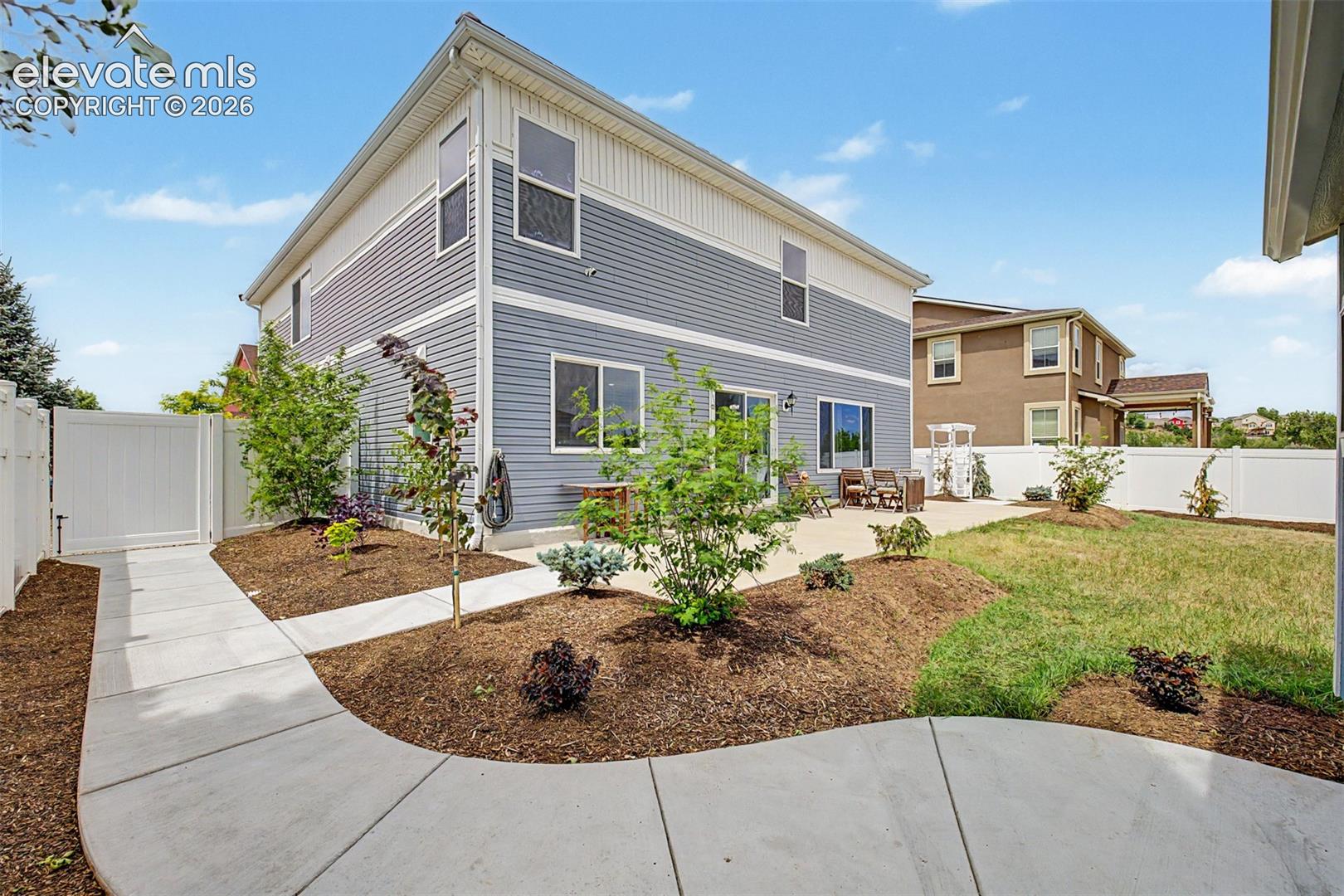 A wide concrete walkway guides you through the lushly landscaped side yard toward the backyard, where young trees and mulched beds frame the path beautifully.