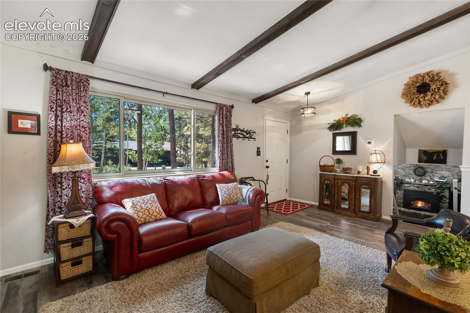 Wood-finish flooring, exposed ceiling beams, a stone fireplace, large windows, and a built-in console