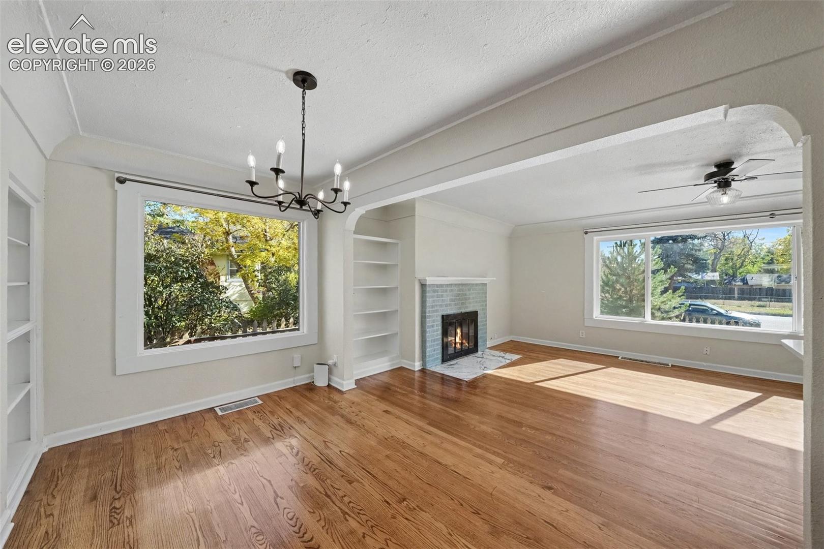 Living area featuring wood-finish flooring, a painted brick fireplace, built-in shelving, a decorative chandelier, and large windows
