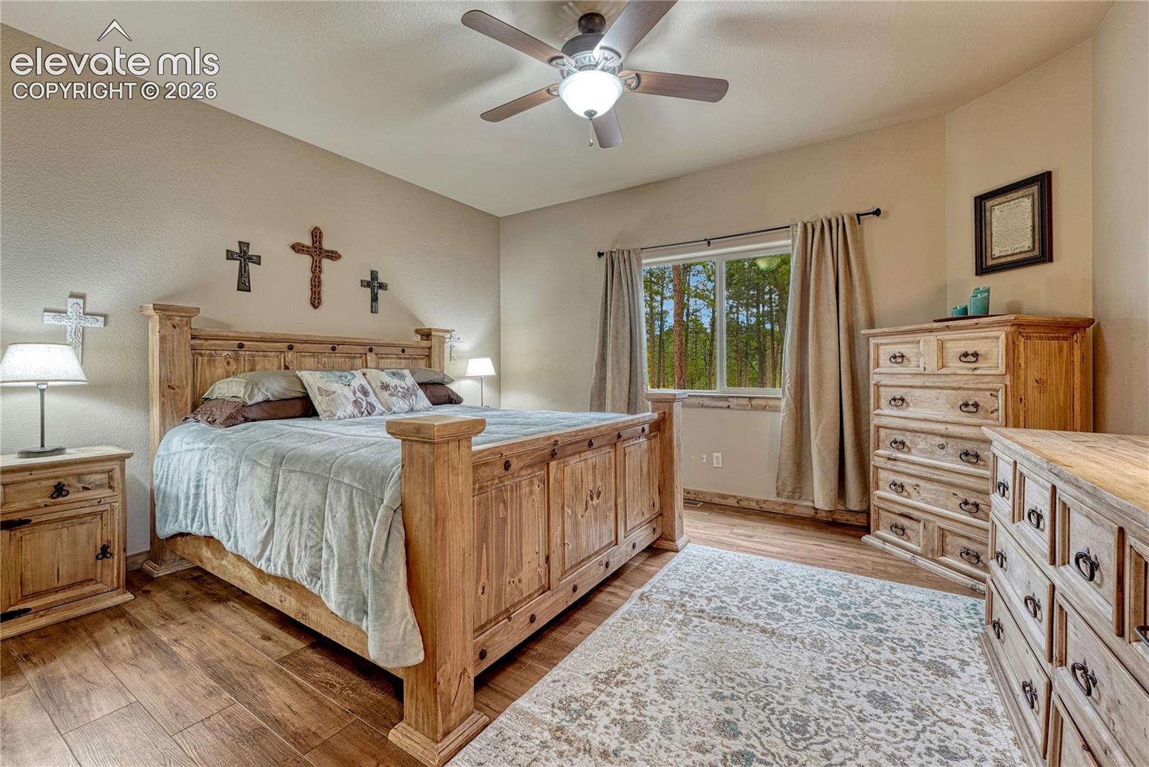 Wood-finish flooring, ceiling fan with integrated lighting, and window featuring an exterior forest backdrop