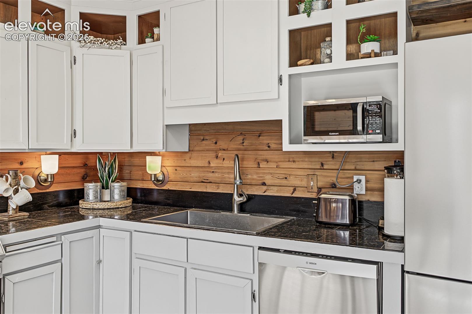White cabinetry, wood-finish backsplash, dark countertops, undermount sink, and stainless steel dishwasher
