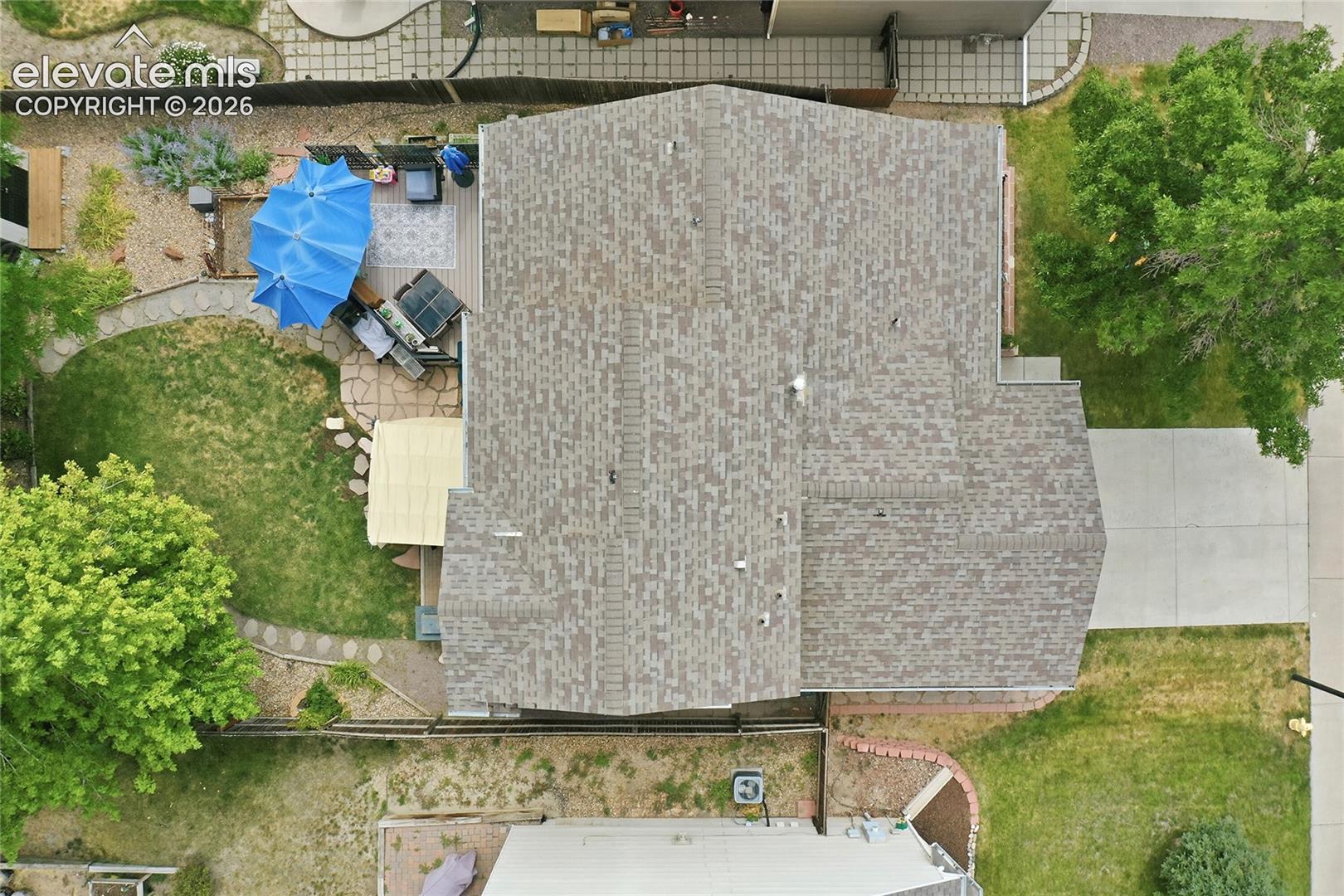Aerial perspective of the property featuring a composition shingle roof, a concrete driveway, a rear patio with stone-finish pavers, and mature landscaping