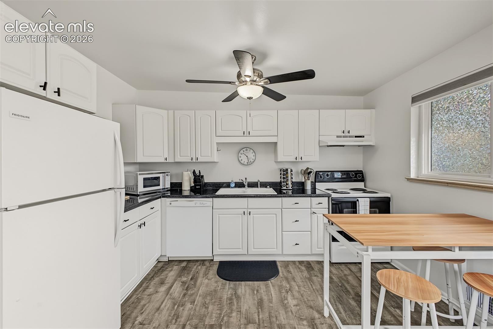 Kitchen featuring white cabinetry, dark countertops, white dual-basin sink, wood-finish flooring, and a ceiling-mounted light fixture with fan