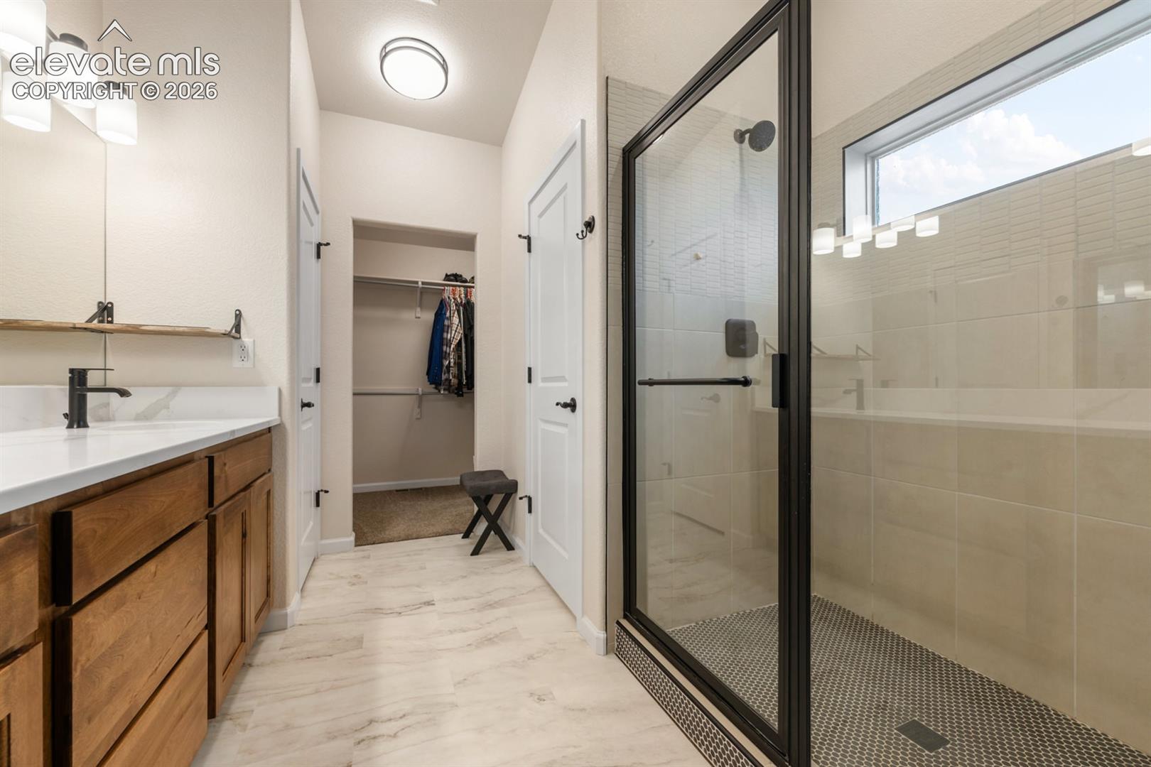 Bathroom featuring a wood-finish vanity with a light countertop, a glass-enclosed shower with tiled walls and flooring, and a walk-in closet with built-in shelving