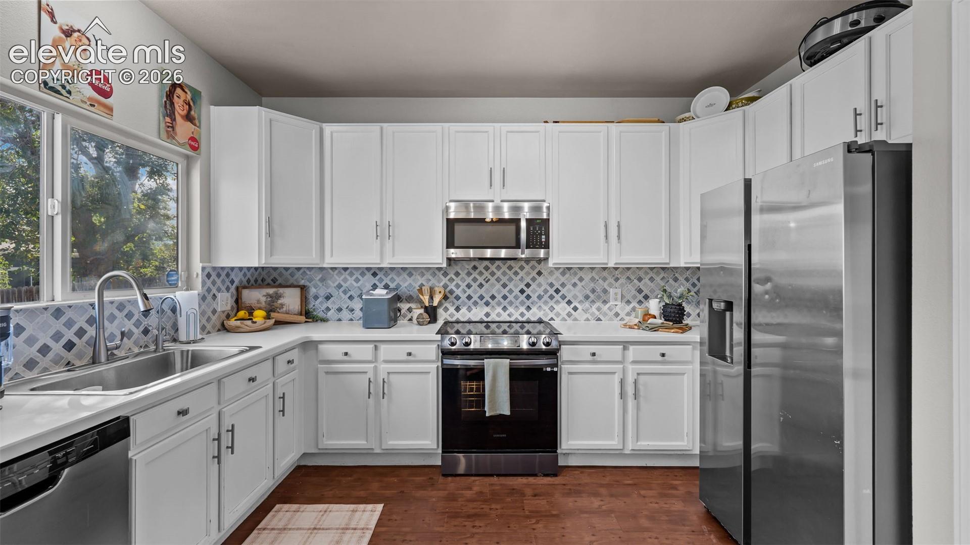 White cabinetry with matte black hardware, stainless steel appliances, patterned tile backsplash, wood-finish flooring, and a large window