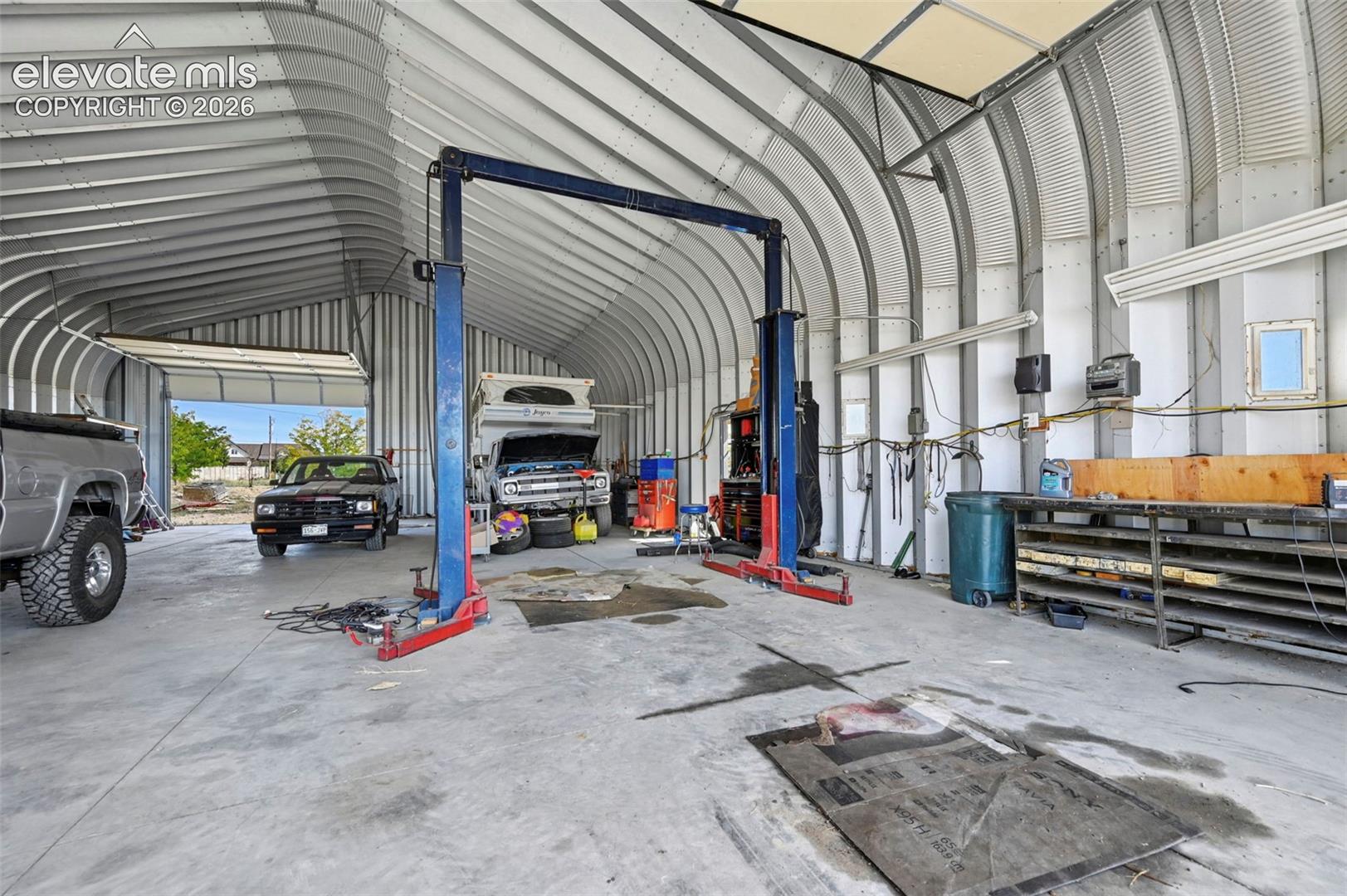 Quonset-style structure featuring a high-clearance vaulted ceiling, industrial concrete flooring, wall-mounted steel shelving, and a professional-grade automotive lift