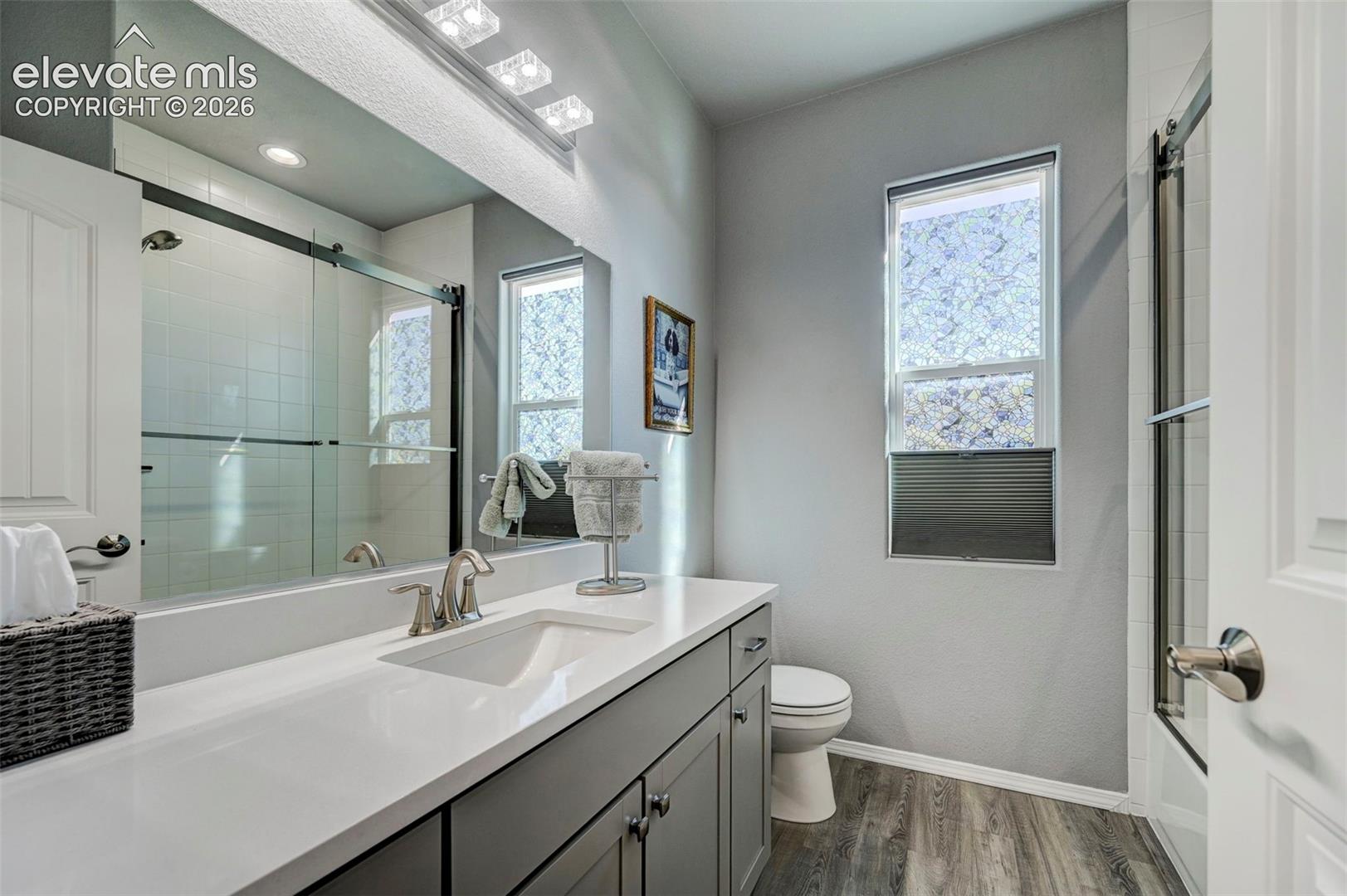 Bathroom featuring a spacious vanity with an undermount sink and a white countertop, dark wood-finish cabinetry, a large frameless mirror, a glass-enclosed shower with subway tile, and wood-finish flooring