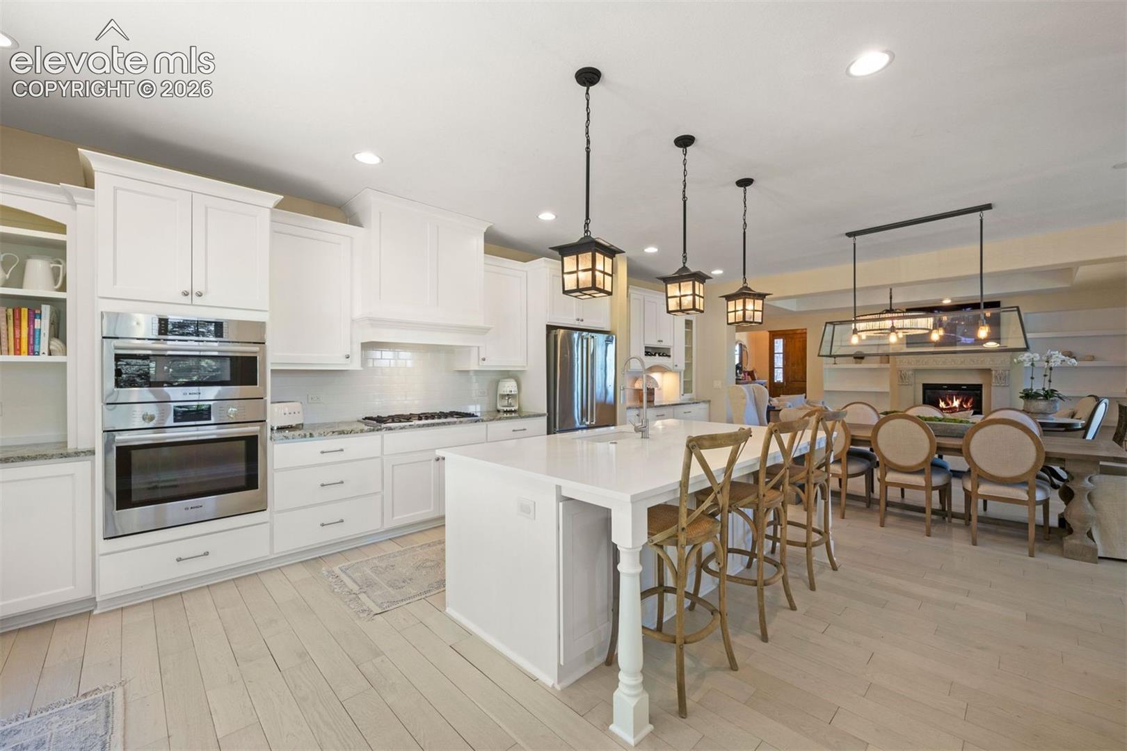 Kitchen featuring white shaker cabinetry, stainless steel double ovens, a built-in range hood, light wood-finish flooring, and a central island with integrated seating