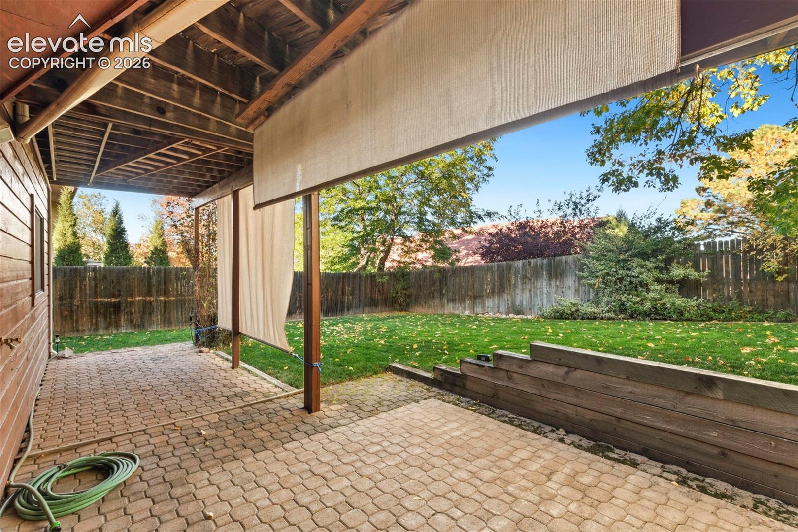 Covered patio featuring paver flooring, timber support posts, and a retractable privacy screen, complemented by a backyard with a manicured lawn and tiered wood retaining wall