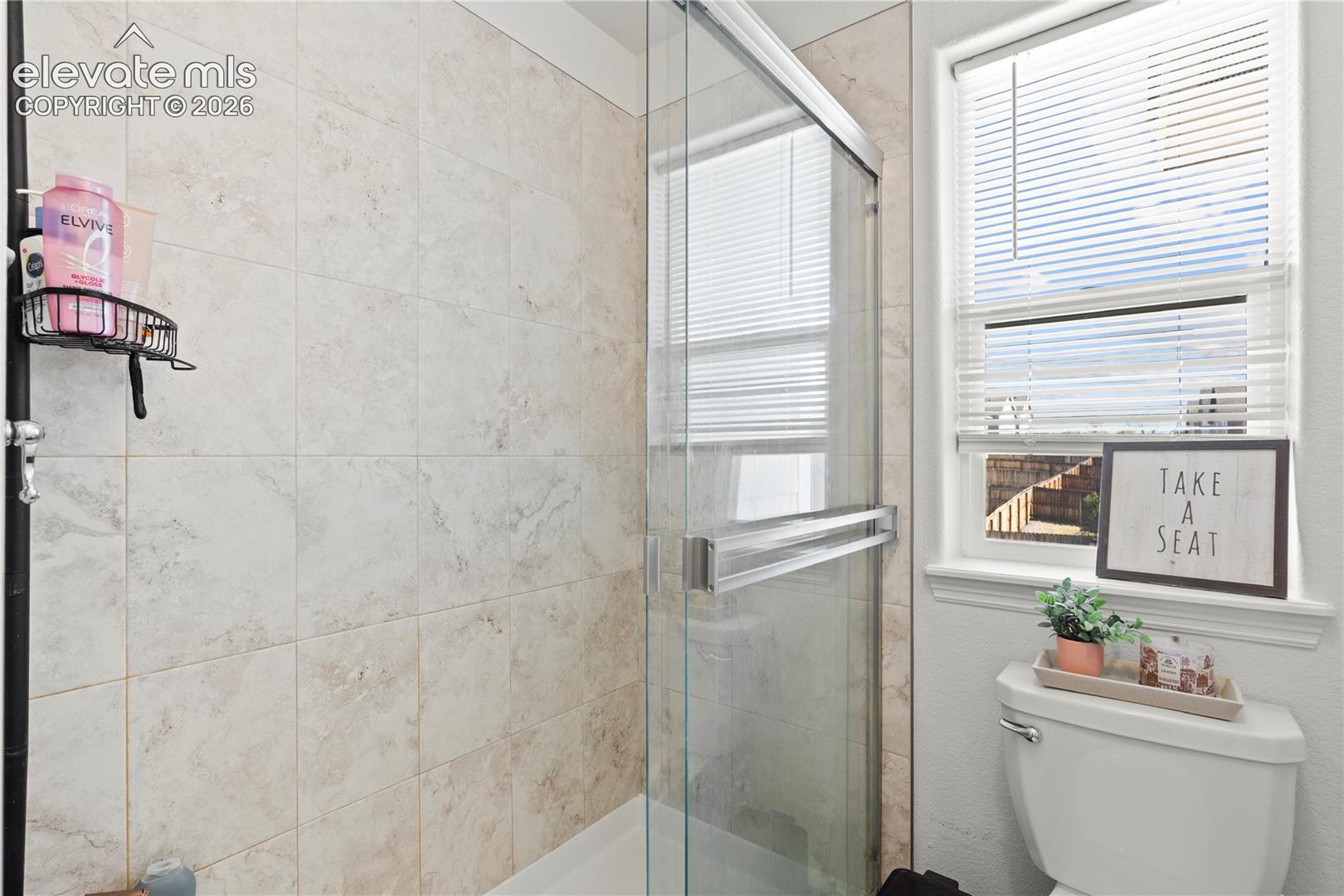 Full-height tiled shower surround, clear glass sliding shower door, polished metal hardware, and a window with horizontal blinds