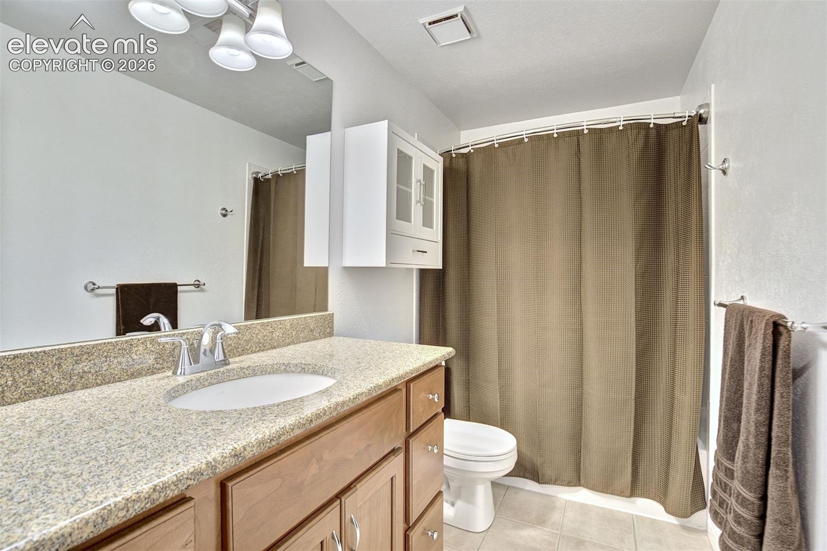 Bathroom featuring a stone-composite countertop, undermount sink with dual-handle polished chrome faucet, wood cabinetry, wall-mounted storage unit with glass-panel doors, and integrated lighting fixtures