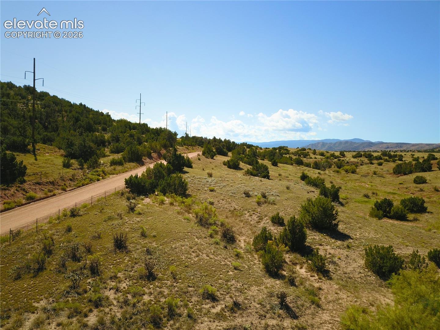 Expansive rural landscape featuring a winding dirt road, scattered green shrubs, rolling hills, and distant mountains