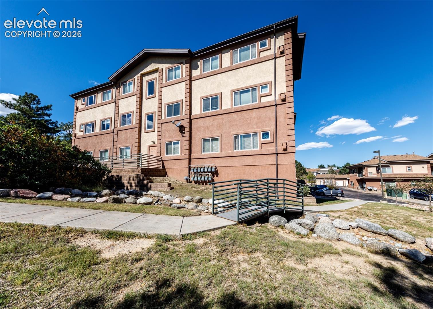 Multi-story exterior featuring a two-tone stucco facade, horizontal metal railings, concrete walkways, and decorative landscape rock bordering