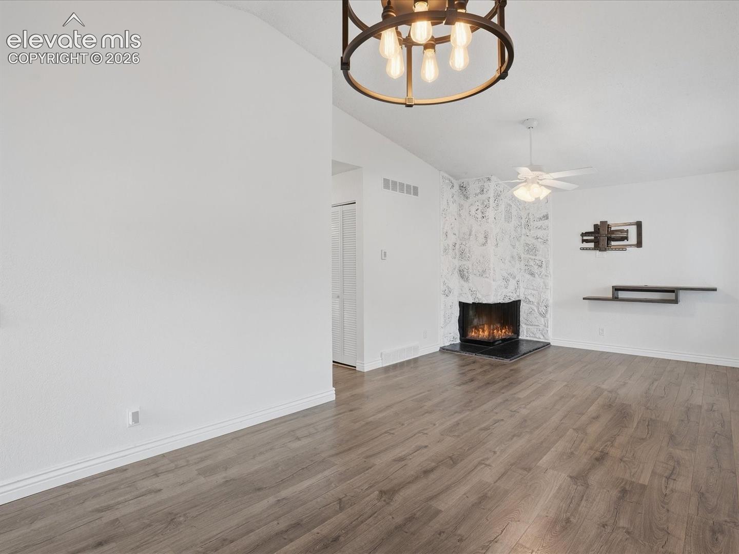 Vaulted ceilings, wood-finish flooring, stone-faced fireplace with a black hearth, industrial-style drum chandelier, and integrated wall-mounted shelving
