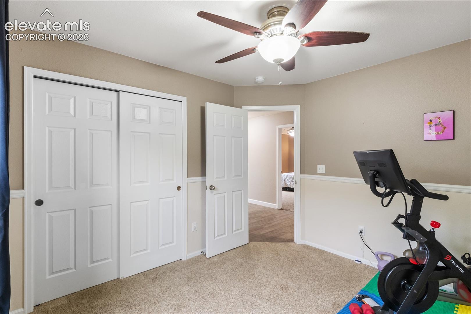 Room featuring wall-to-wall carpeting, white dual-panel closet doors, white paneled interior door, chair rail molding, and a ceiling fan with integrated lighting