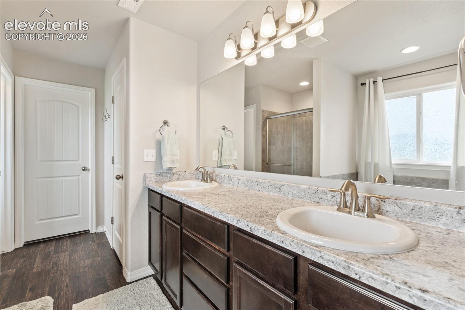 Dual vanity with undermount sinks and extensive countertop space, large wall-mounted mirror, brushed nickel fixtures, and wood-finish flooring