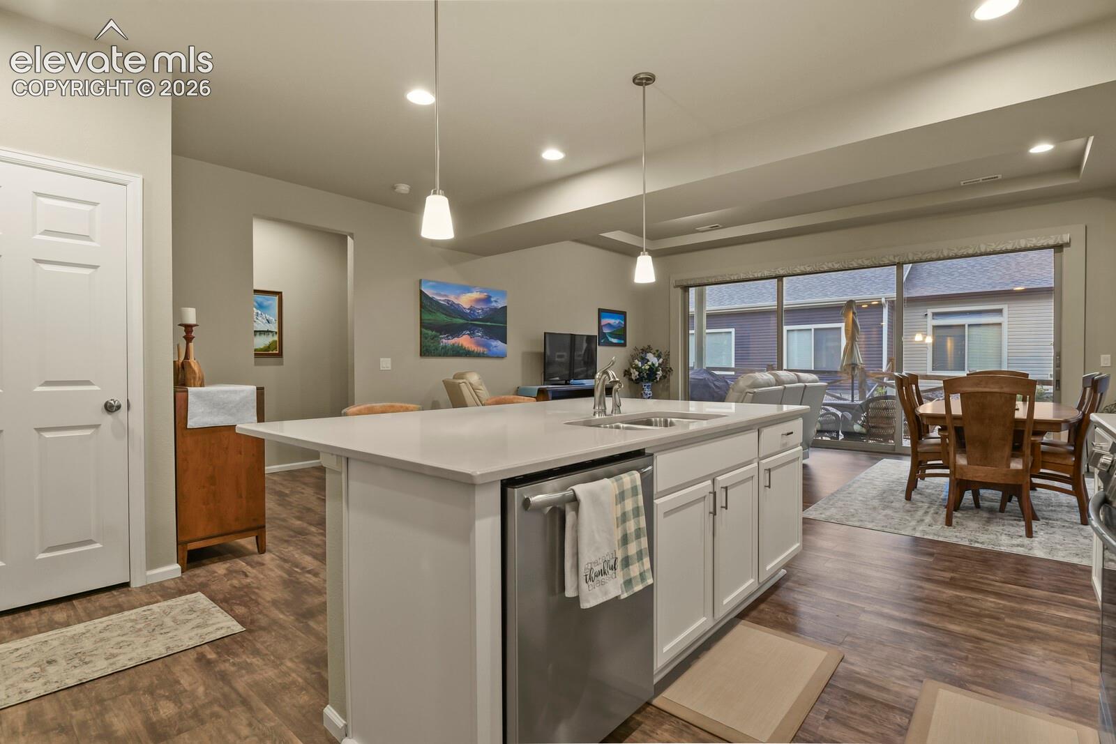 Kitchen island featuring white cabinetry, a light-toned solid surface countertop, stainless steel dishwasher, and dual pendant lighting, complemented by wood-finish luxury tile flooring