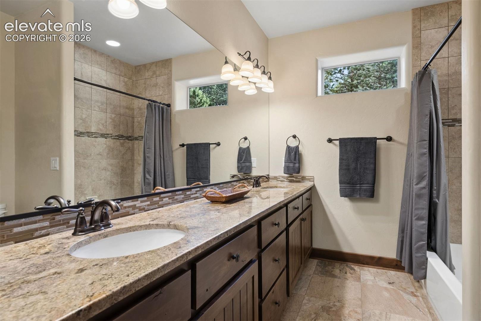 Dual-sink vanity with stone-finish countertop, full-width mirror, tile-surround shower, and tile flooring