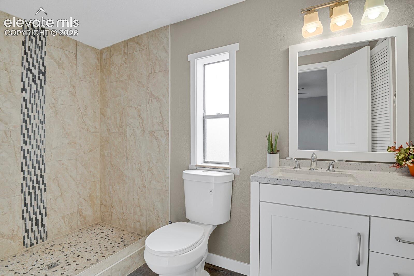 Walk-in shower with natural stone-look tiling and mosaic accent, pebble shower floor, single vanity with integrated sink and stone-finish countertop, and wall-mounted light fixture