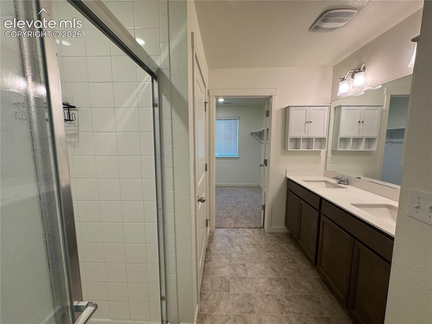 Bathroom featuring a dual-sink vanity with light-colored solid surface countertops, dark-finish cabinetry, a glass-enclosed shower with white wall tile, and wall-mounted white storage cabinetry
