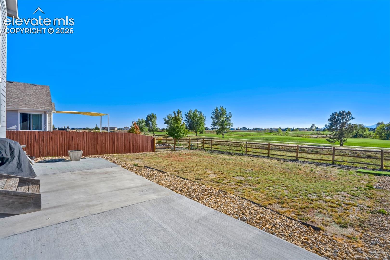 Expansive concrete patio with a gravel border, wood privacy fence, and a distant field beyond a split-rail fence