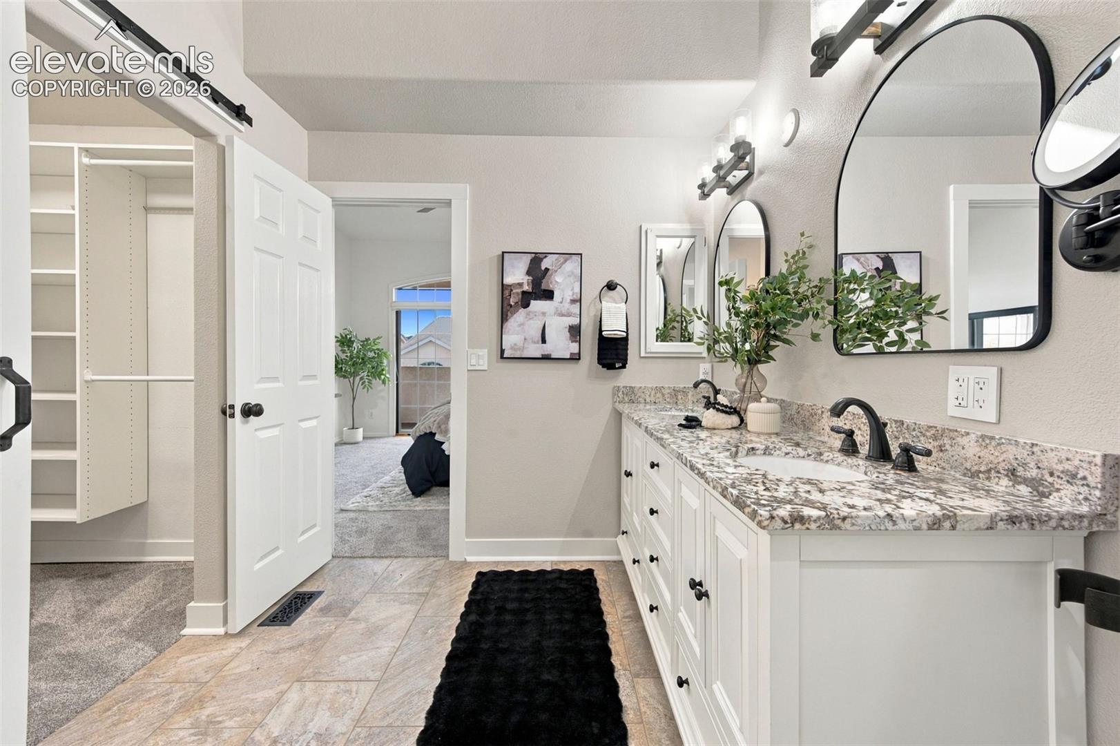 Double vanity featuring white cabinetry, patterned stone countertop, dual undermount sinks, and matte black hardware with arched wall-mounted mirrors and custom built-in closet shelving