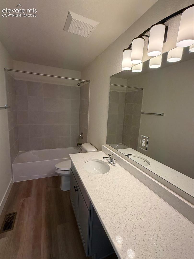 Bathroom featuring a long vanity with a solid surface countertop, an integrated sink, full-width wall mirror, five-light fixture, and tub-shower combination with tiled surround, finished with wood-finish flooring
