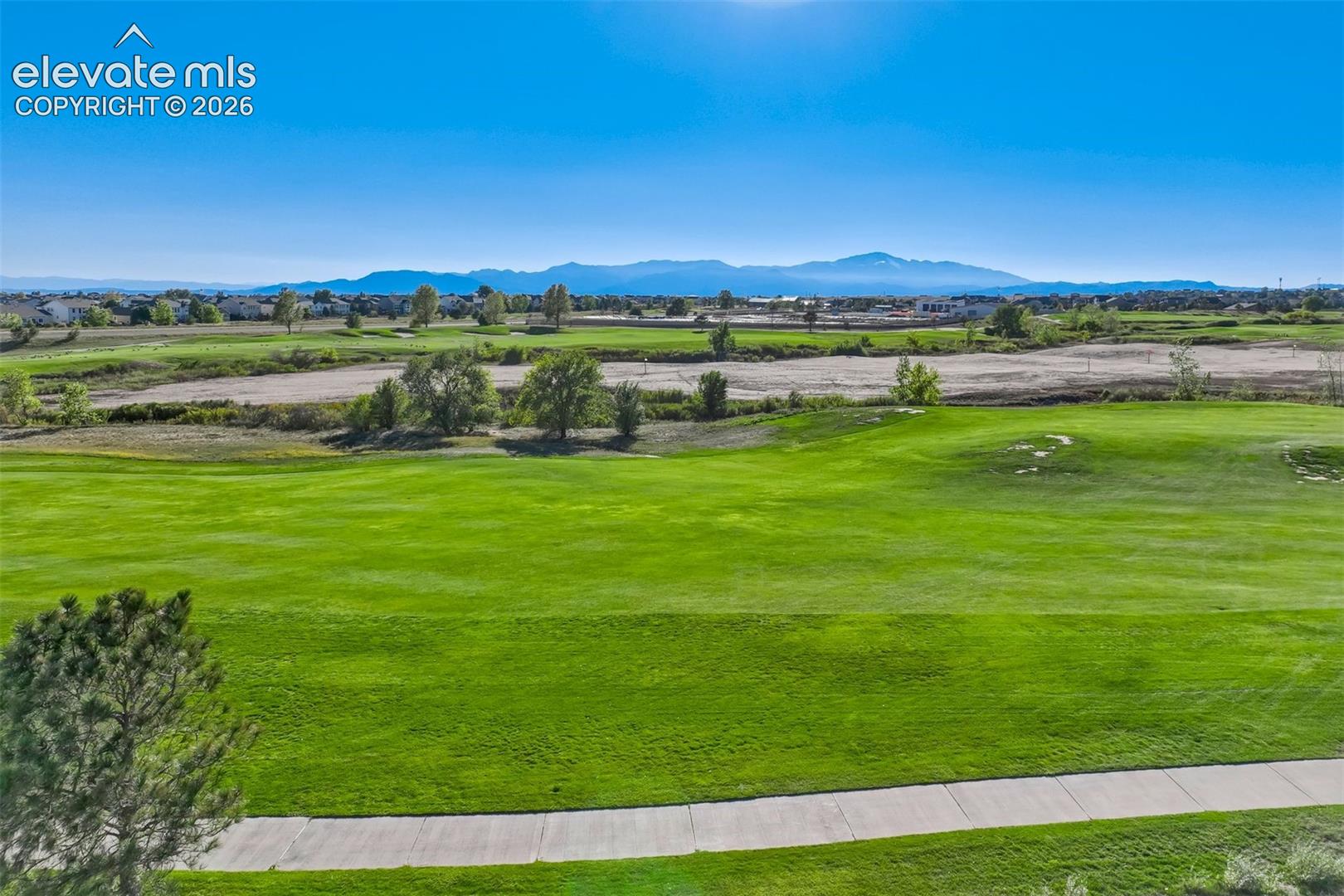 Expansive green golf course with a paved path, surrounded by distant residential structures and mountains