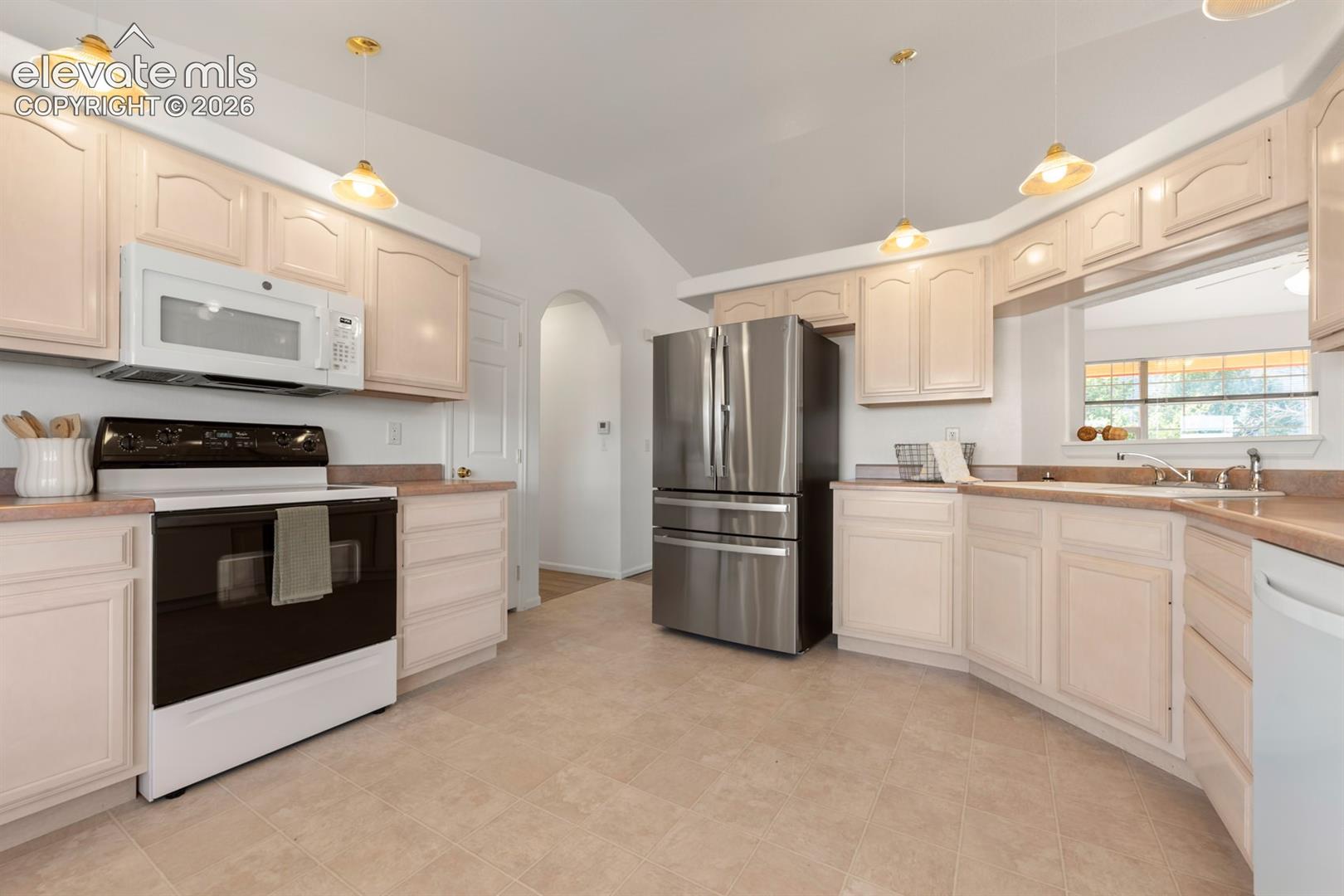Kitchen featuring light-toned cabinetry, stainless steel refrigerator, arched doorway, vaulted ceiling, and pendant lighting