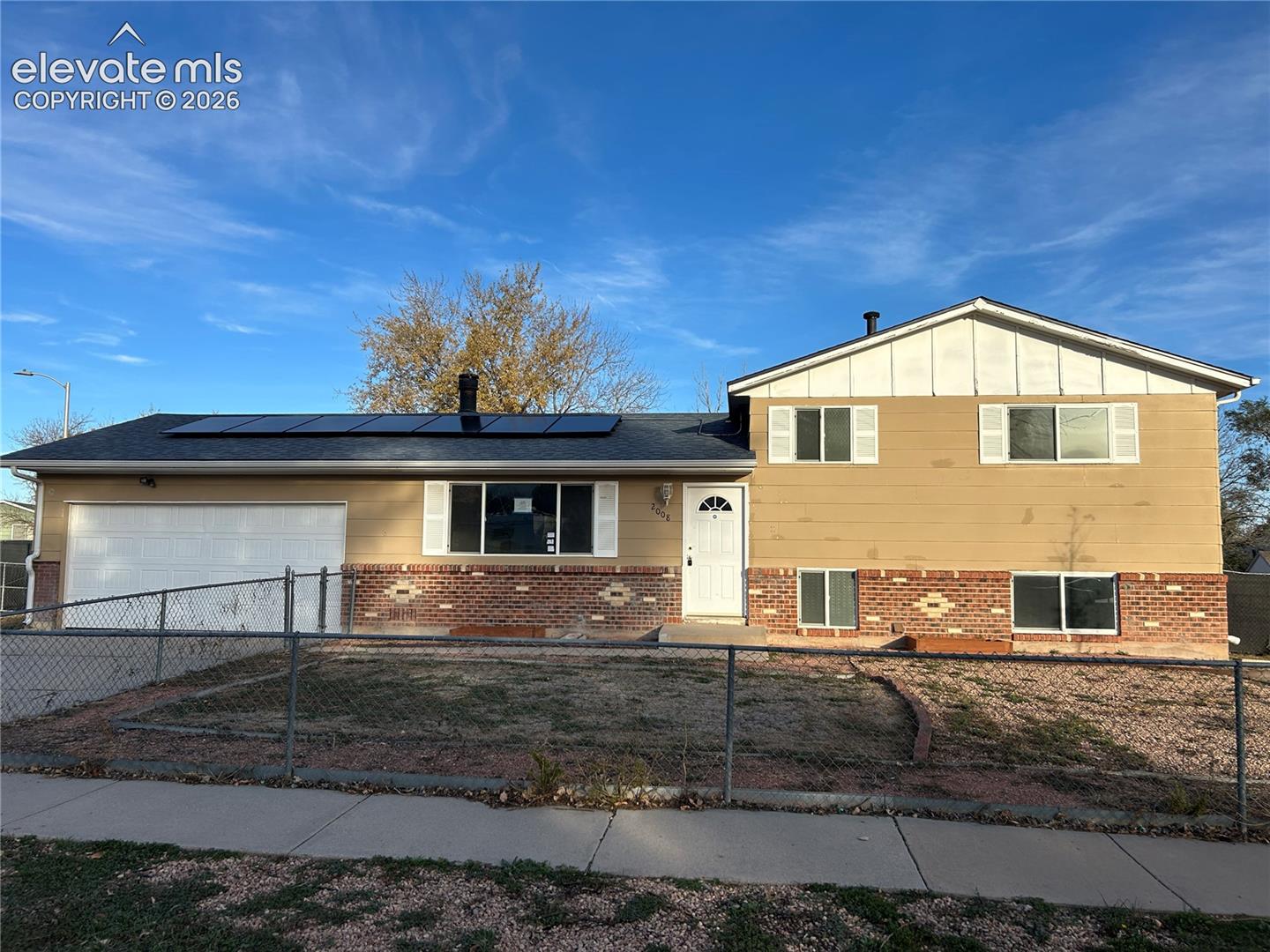 Split-level exterior featuring a brick and horizontal siding facade, roof-mounted solar panels, an attached two-car garage, and chain-link fencing