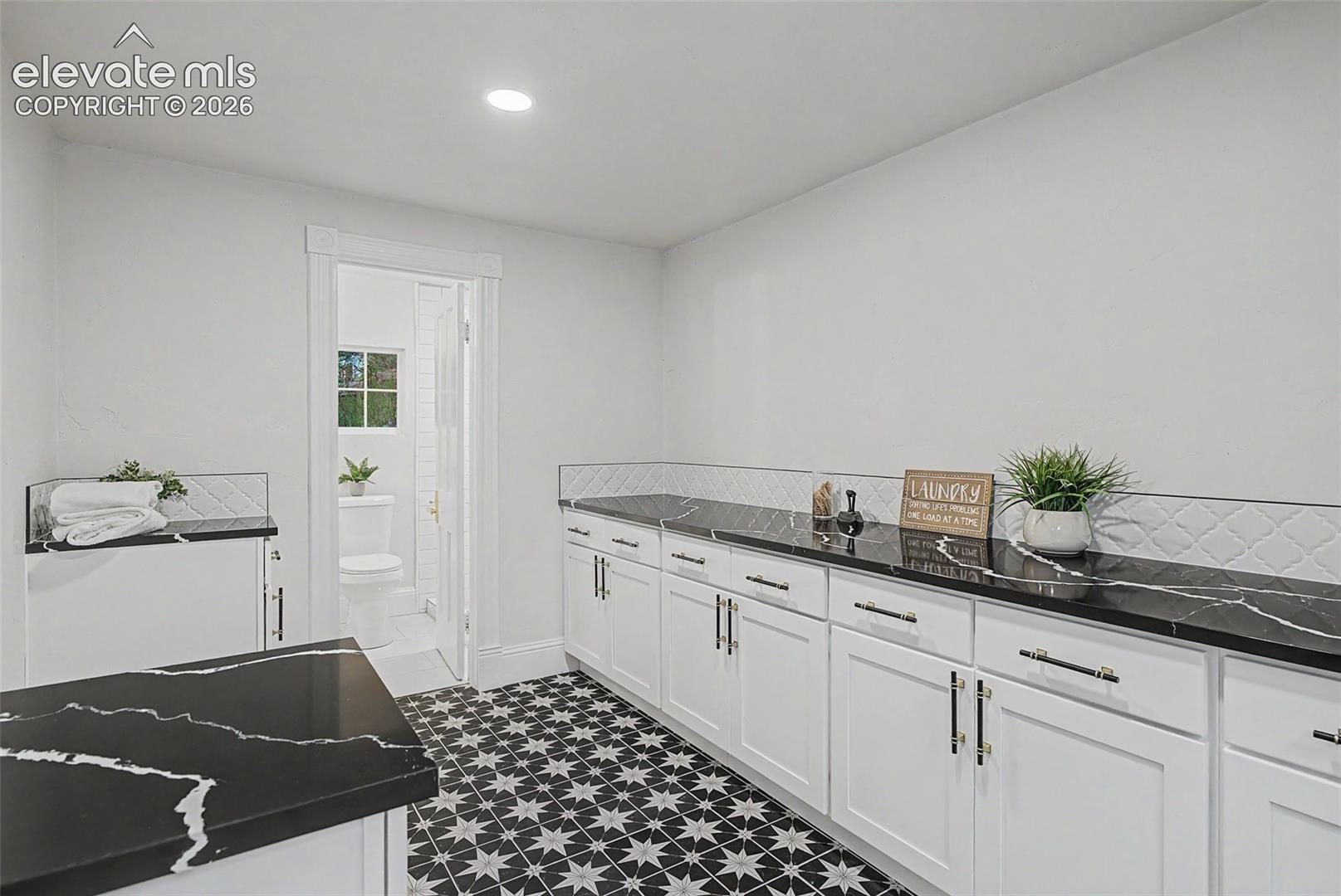 Spacious utility room featuring patterned tile flooring, extensive black countertops with white veining, white cabinetry with black hardware, and a decorative tile backsplash