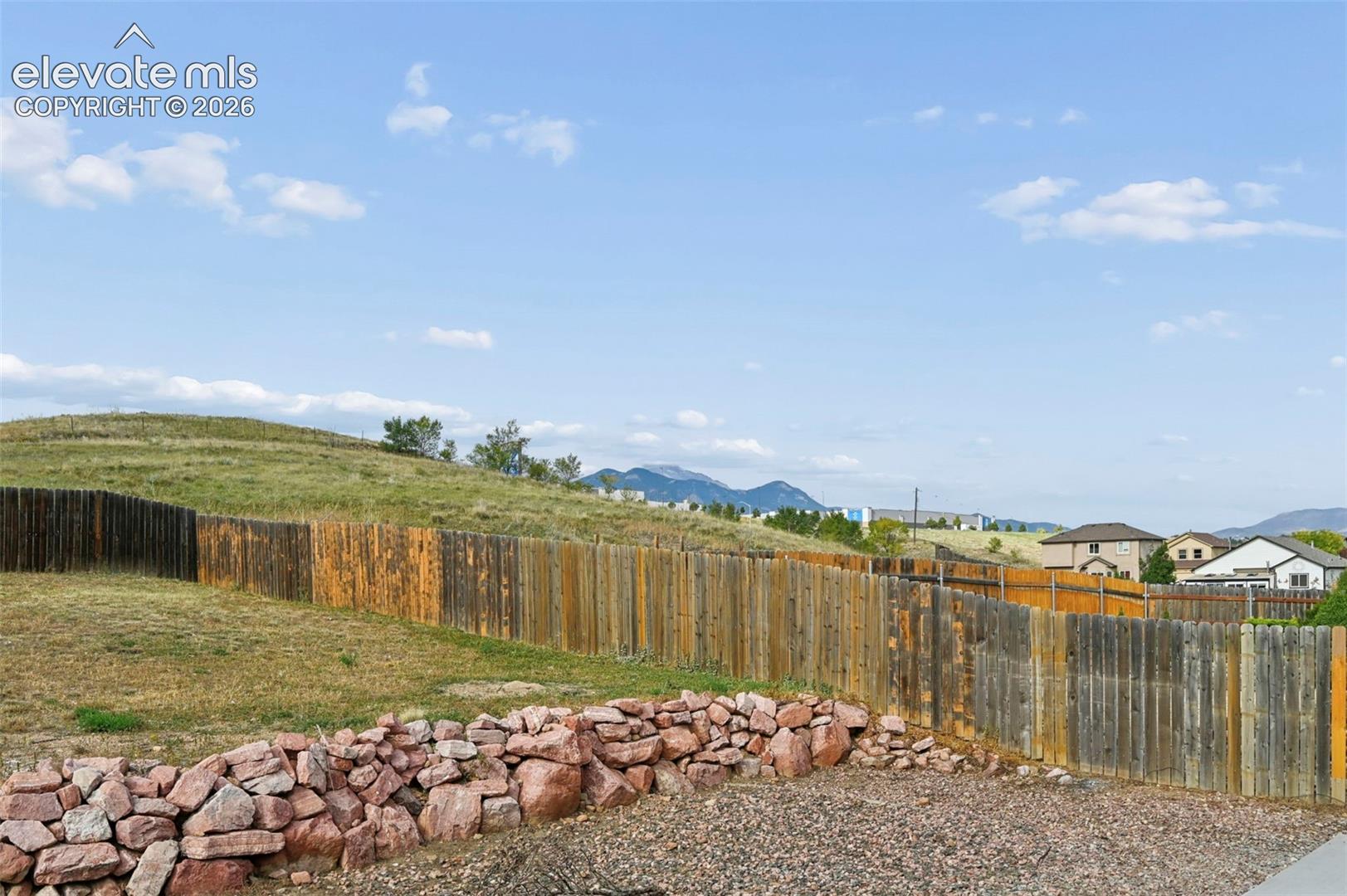 Fenced backyard featuring natural stone retaining walls, gravel landscaping, concrete patio sections, and mountain backdrop