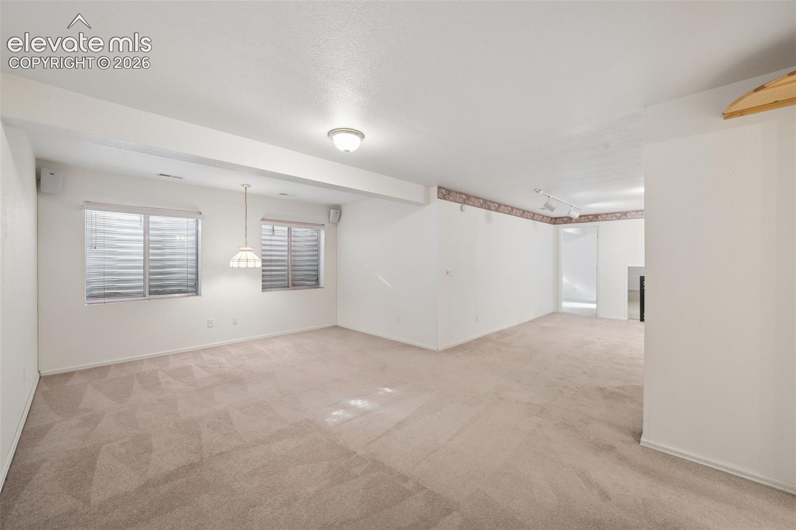 Open-concept interior featuring neutral wall-to-wall carpeting, dual windows with horizontal blinds, a ceiling-mounted pendant light, and integrated track lighting