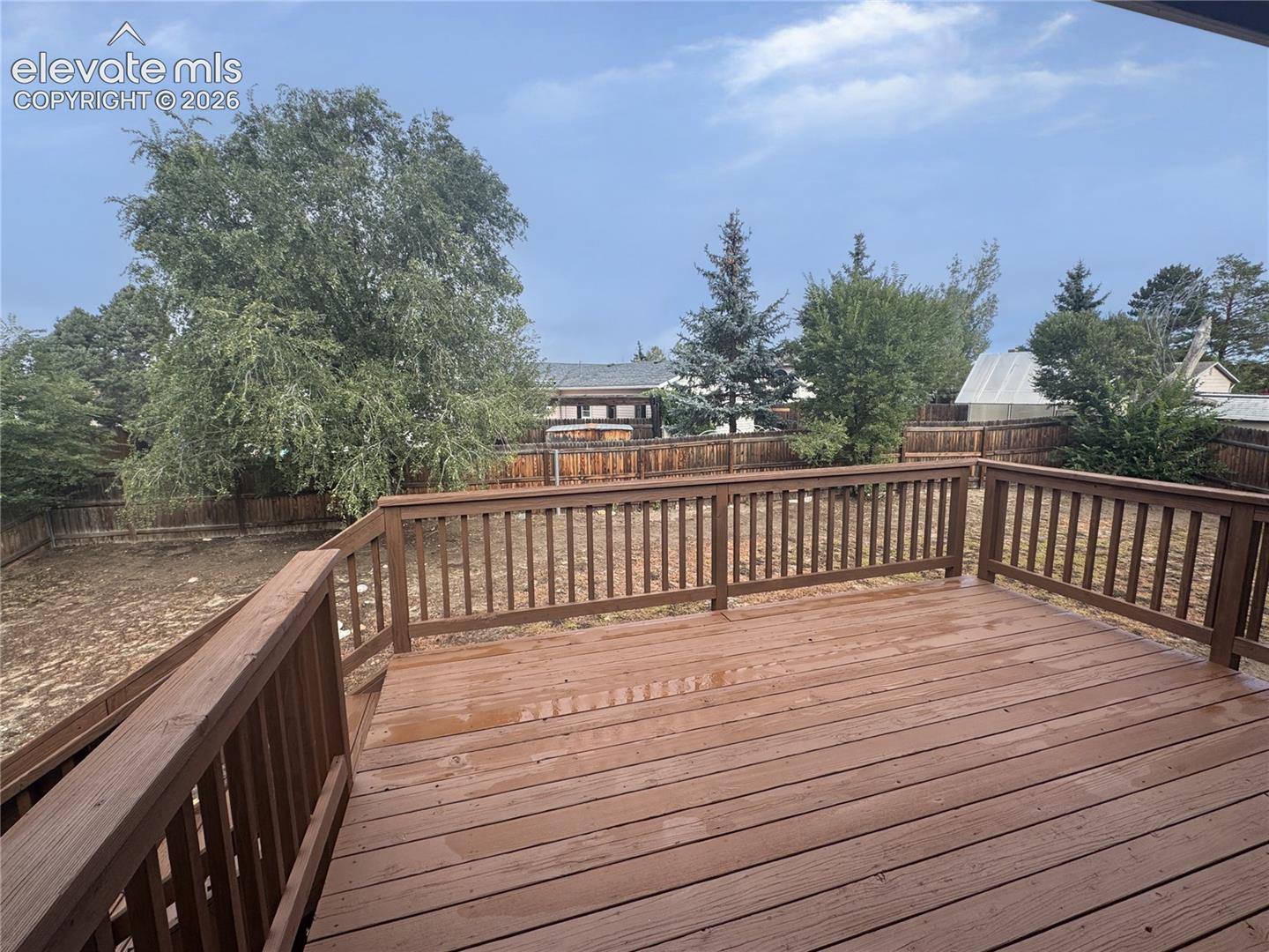 Elevated wood deck featuring vertical baluster railings, enclosing a spacious yard with mature trees and a full-perimeter wood privacy fence