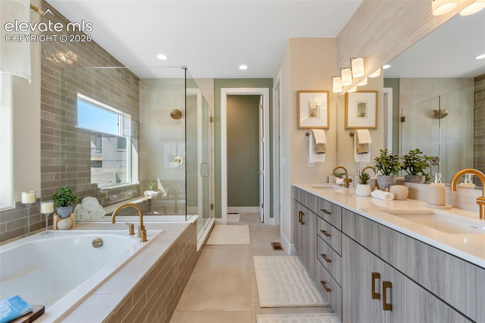 Bathroom featuring a double-sink vanity with light wood-finish cabinetry, a white soaking tub with metallic fixtures, a glass-enclosed walk-in shower, tile surround accents, and a full-width wall mirror