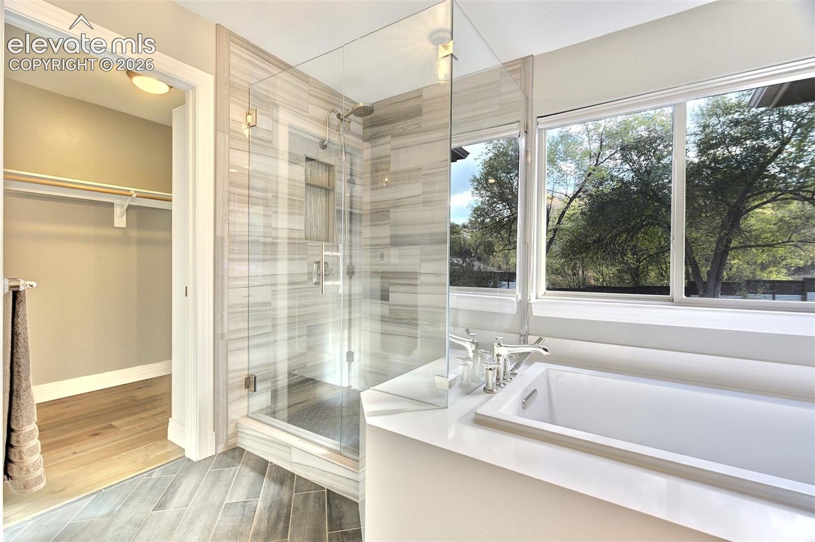 Glass-enclosed walk-in shower with vertical wall tile and built-in niche, soaking tub featuring a wide ledge, wood-finish flooring, and horizontal picture windows