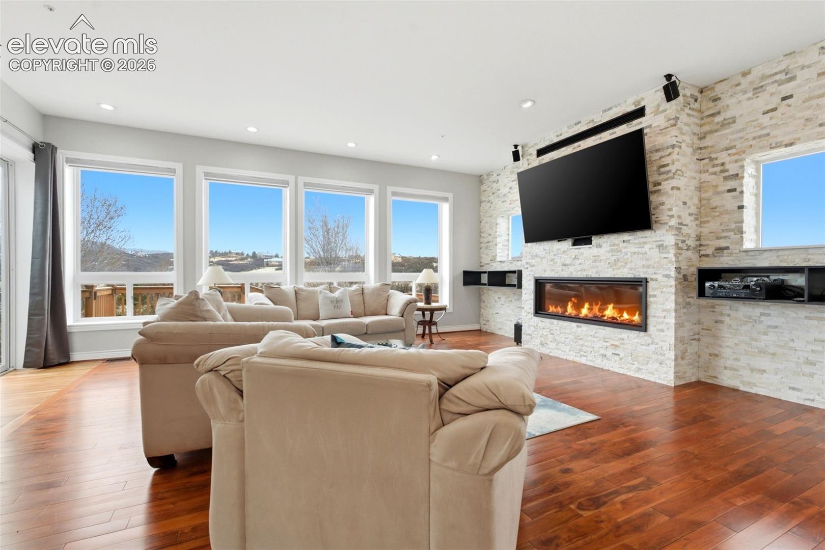Hardwood flooring, linear fireplace with stone surround, wall-mounted television, multiple windows, and recessed lighting