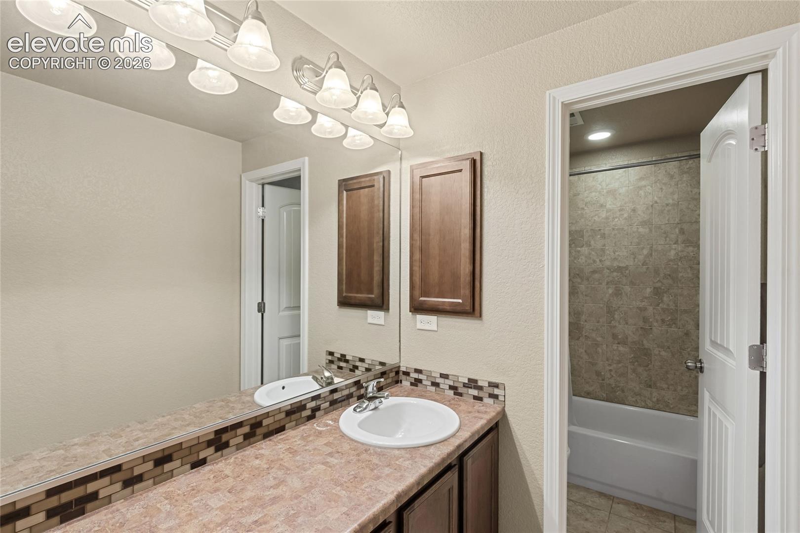 Bathroom featuring a solid-surface countertop, integrated vanity sink, decorative mosaic tile backsplash, wood-finish cabinetry, and a full-width wall mirror with multi-bulb light fixture