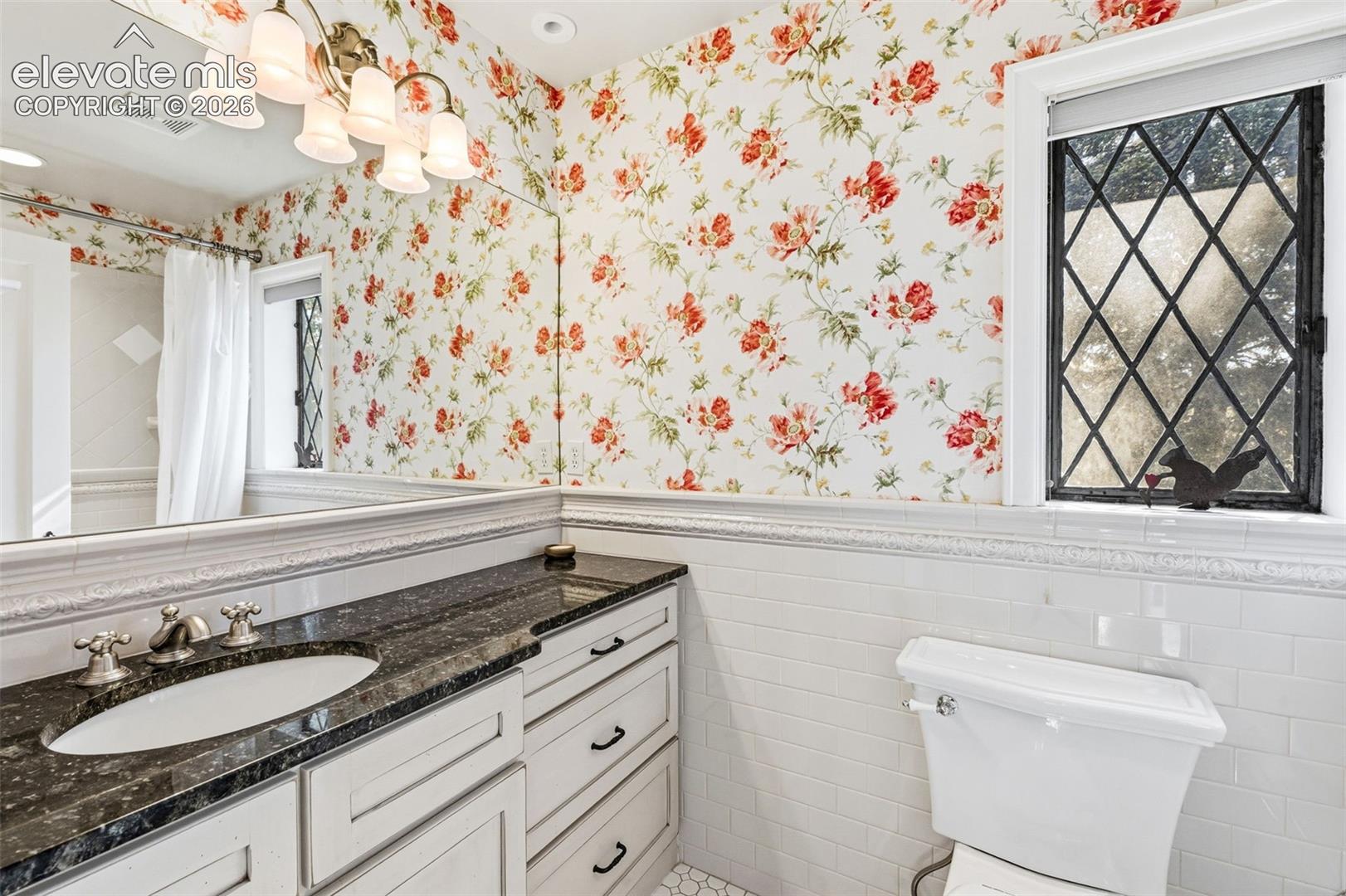 Vanity featuring a dark stone countertop, integrated oval sink, and white cabinetry with matte hardware, accented by classic subway tile wainscoting and a leaded glass window