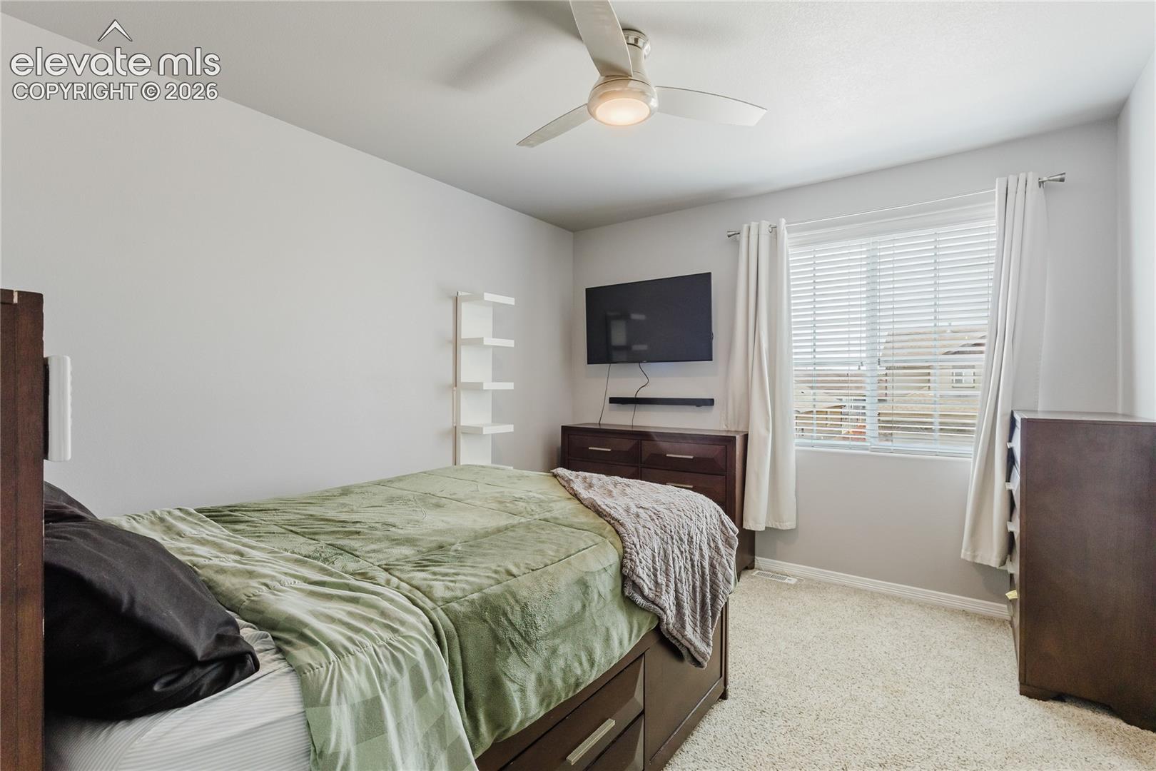 Carpeted room featuring a ceiling fan with integrated lighting, a window with blinds and drapes, and a built-in wall shelf