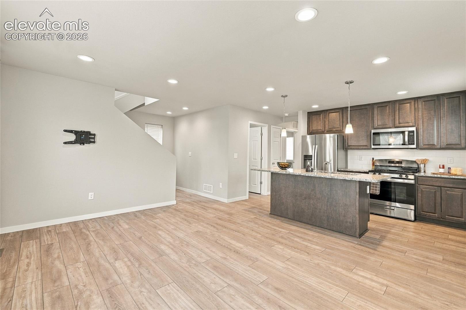 Open-concept space featuring wood-finish flooring, recessed lighting, and a kitchen with stainless steel appliances, dark wood cabinetry, and a central island with a stone-finish countertop
