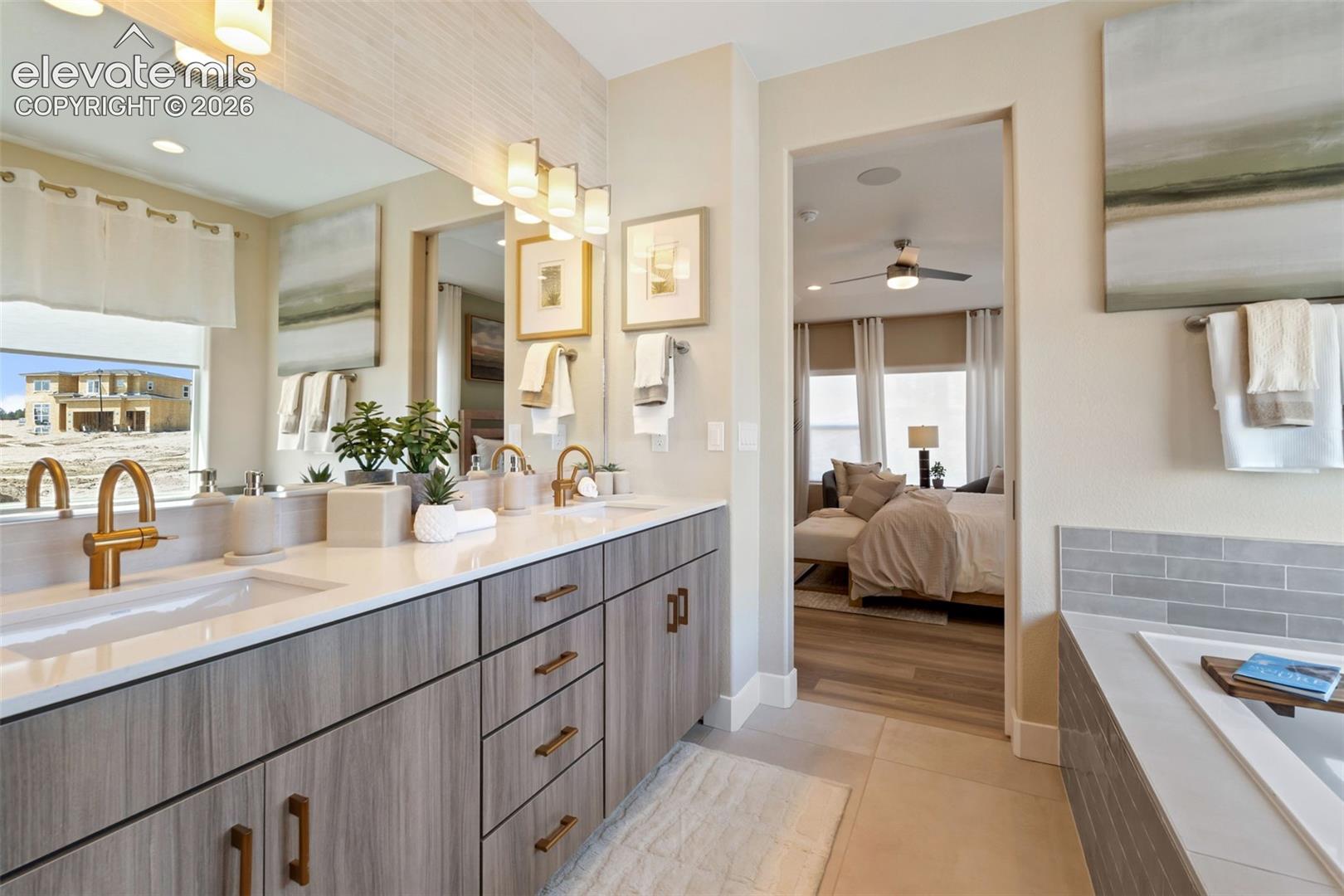 Dual-sink vanity with wood-finish cabinetry, light-toned countertops, brass-toned fixtures, a wall-to-wall mirror, and a soaking tub with tile surround