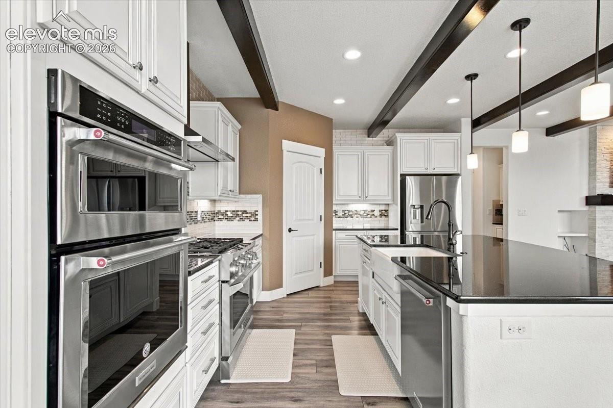 Kitchen featuring shaker-style cabinetry, dark stone countertops, stainless steel double wall ovens, apron-front sink, and exposed ceiling beams