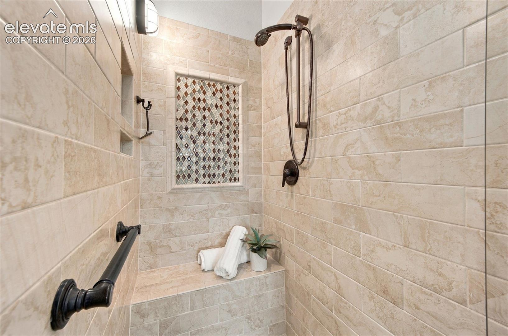 Walk-in shower featuring large-format neutral-tone tile walls, an integrated bench seat, a decorative diamond-pattern tile inlay, an oil-rubbed bronze rainfall showerhead, and a matching grab bar