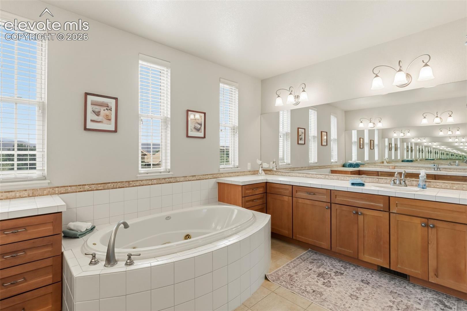 Spacious bathroom featuring an oval JACUZZI soaking tub with tile surround, dual vanities with wood cabinetry, and extensive wall-mounted mirrors