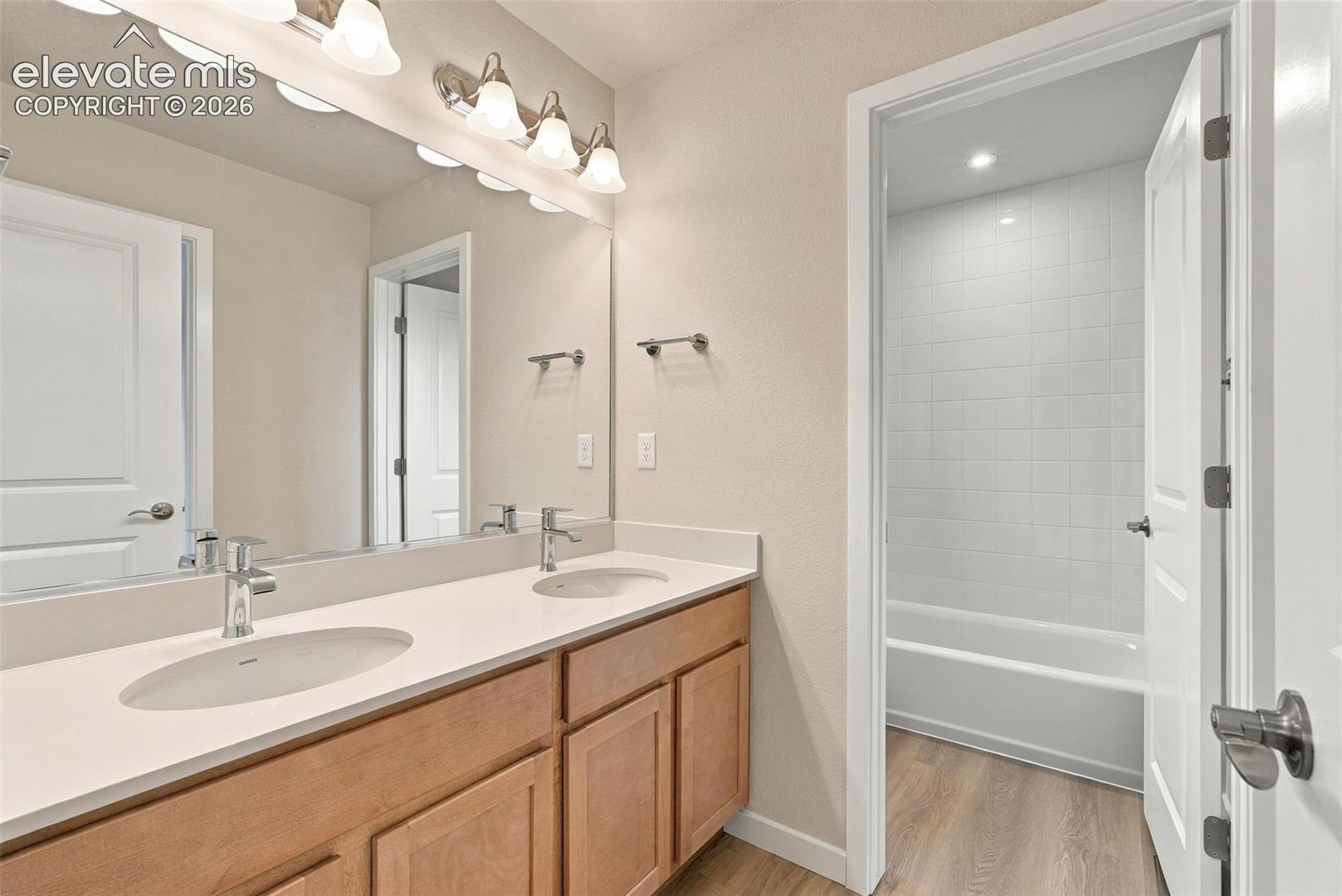 Double vanity featuring wood-finish cabinetry, light-toned countertop with dual integrated sinks, full-width mirror, and a tub-shower combination with tiled surround
